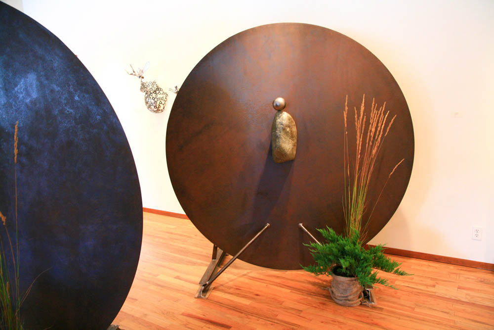 Image of 2 metal sculptures in a gallery a round disc with a rock centered in the middle and a metal ball balanced on top stacy beamer artist